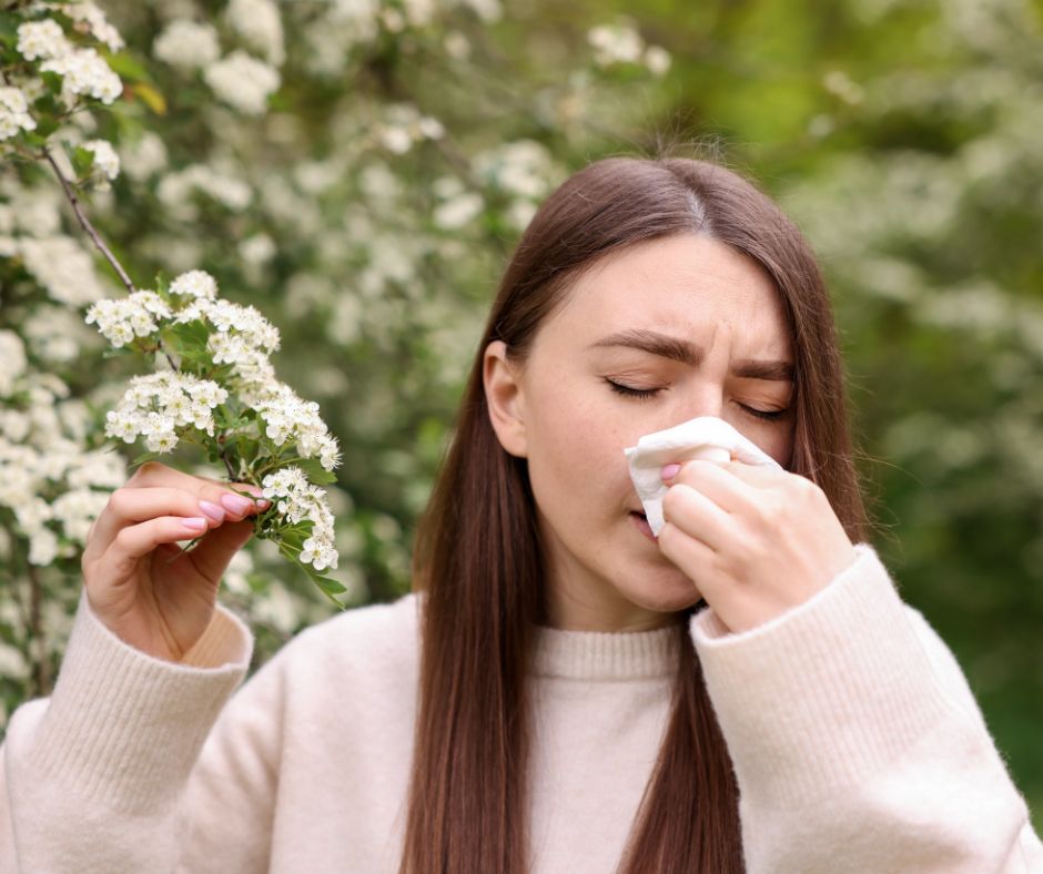 Allergien ganzheitlich behandeln – Junges Mädchen mit Pollenallergie hält eine blähende Pflanze in der Hand, von der sie sich wegdreht und in ein Taschentuch schnäuzt